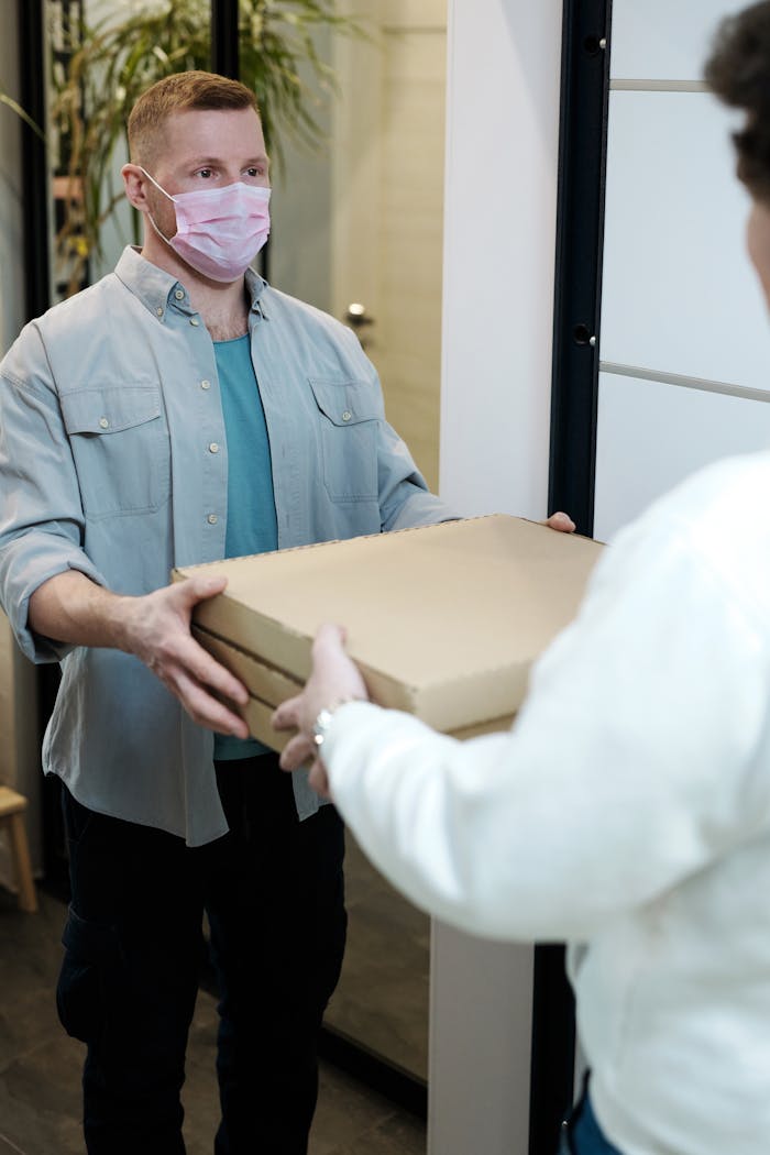 A delivery man with a mask handing over pizza boxes indoors, embodying pandemic safety measures.